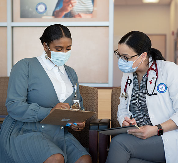 Two women in medical attire with face masks sitting next to each other with note pads looking at one of the note pads