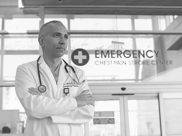 Doctor with his arms crossed, wearing a white lab coat, standing in front of an ER entrance