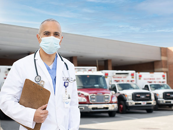 Doctor wearing a face mask and a white lab coat standing in front of ambulances holding a note pad