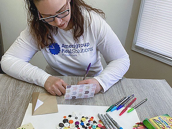 Woman wearing a white long sleeve shirt that says "Amerigroup real solutions" in the front, writing a card with Crayolas, Sharpies, colored pens and buttons spread on a table
