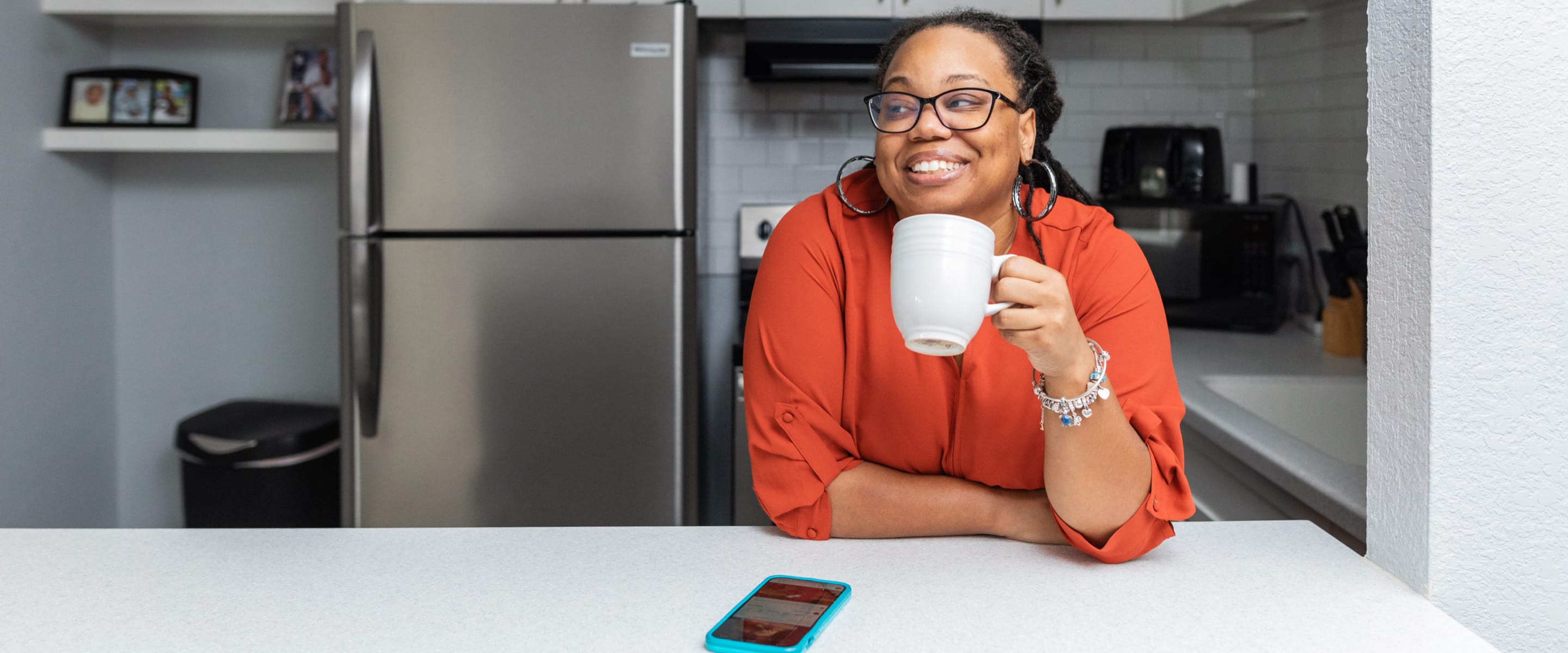 A woman in an orange outfit holding a white mug smiling at a kitchen countertop with a fridge behind her