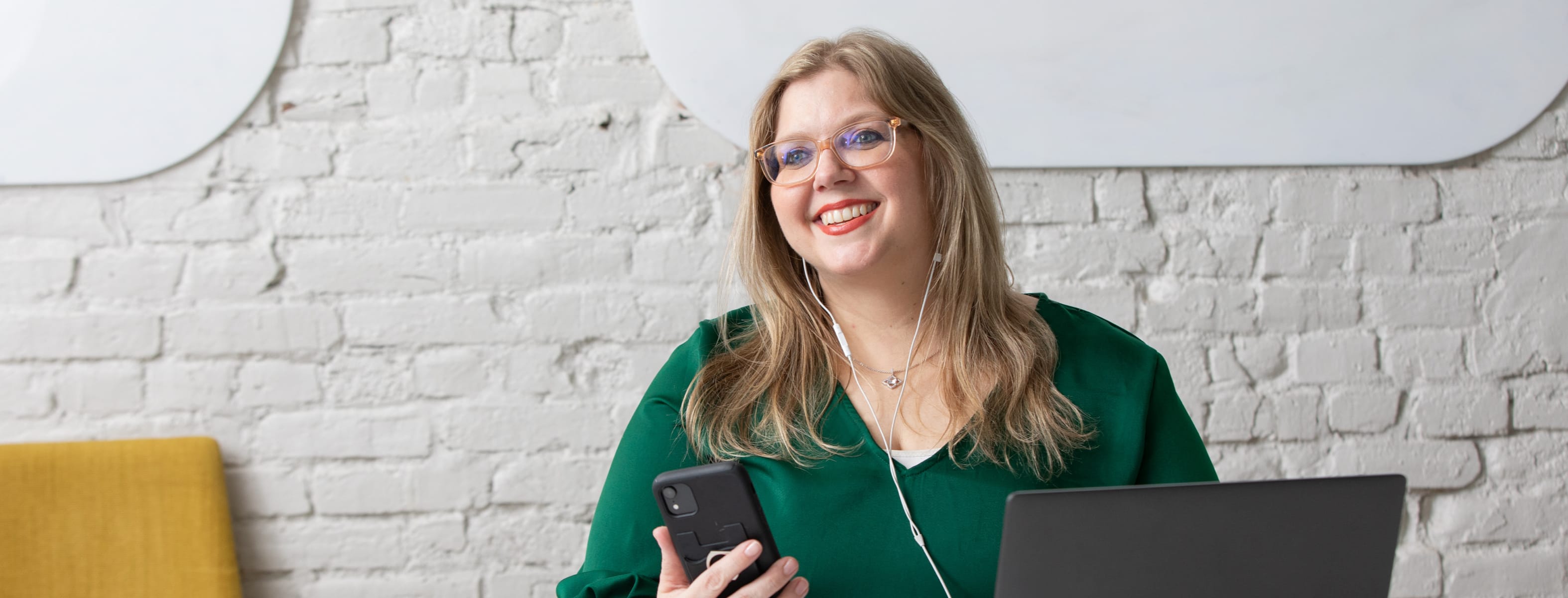A woman in a green outfit with long hair smiling while holding a phone next to a laptop.