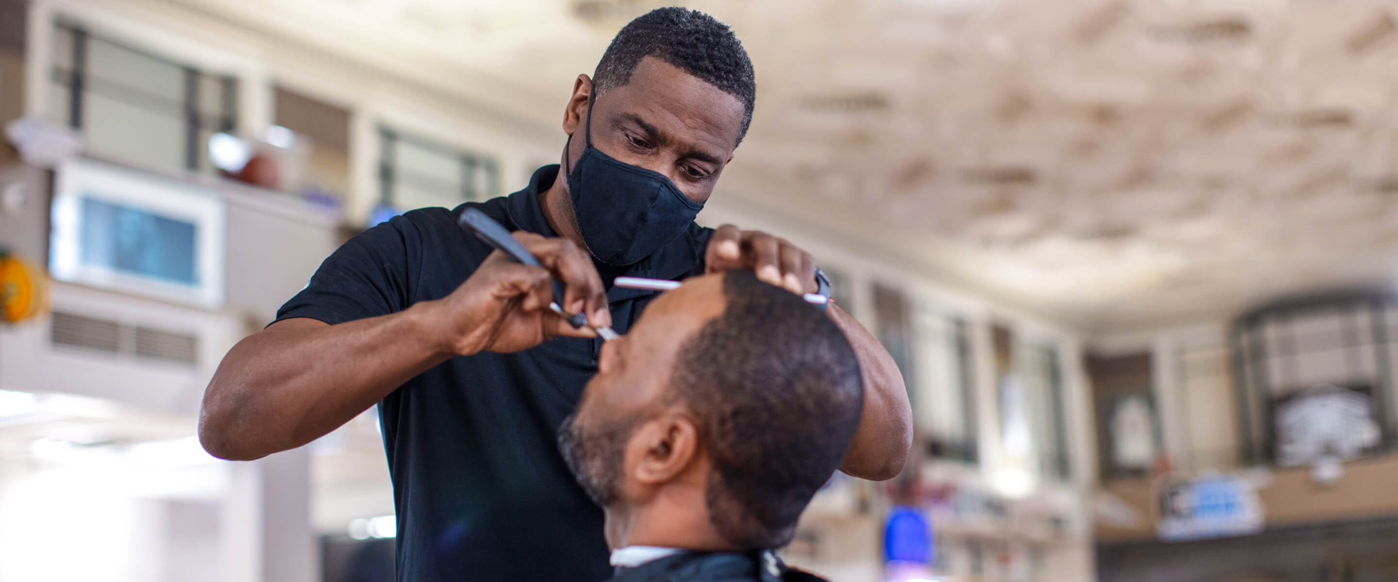 A barber wearing a black mask holding scissors cutting a man's hair.