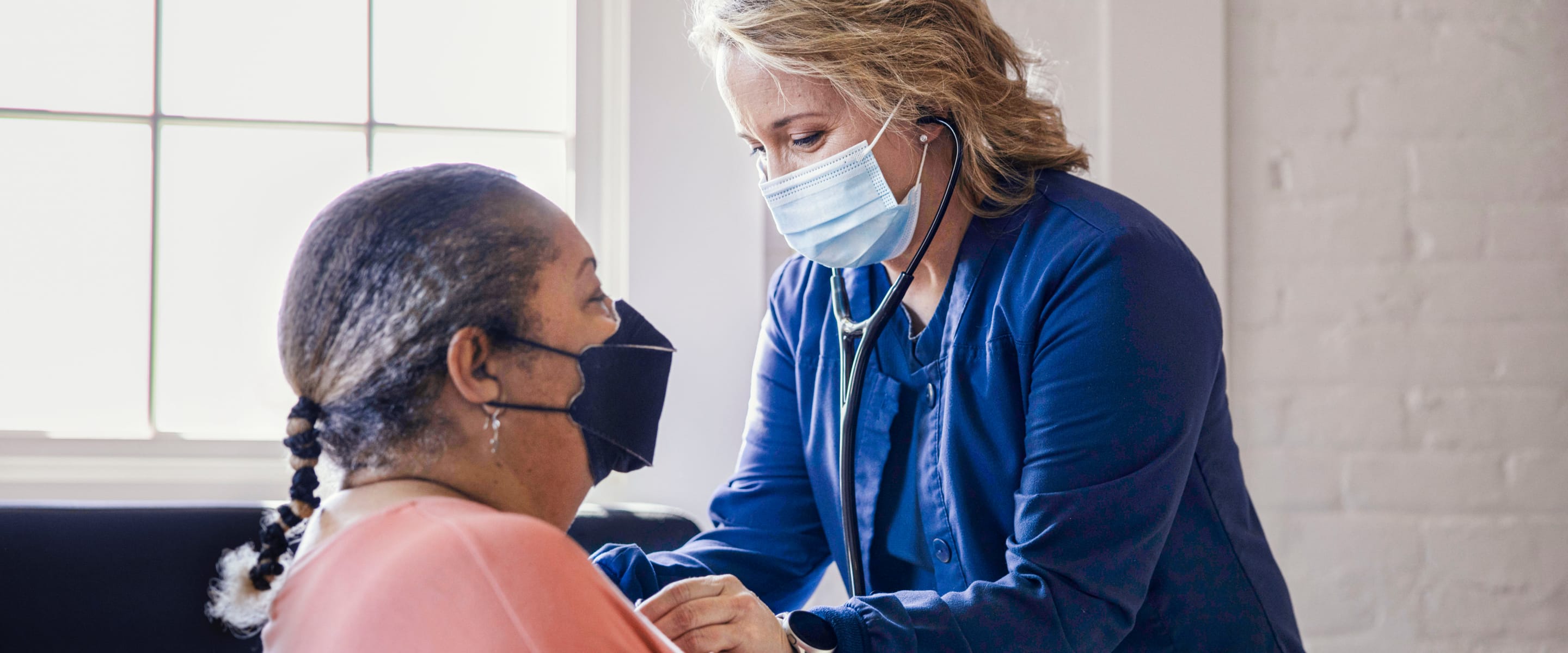 A nurse wearing a mask with a stethoscope leaning in on a woman sitting down with a mask.